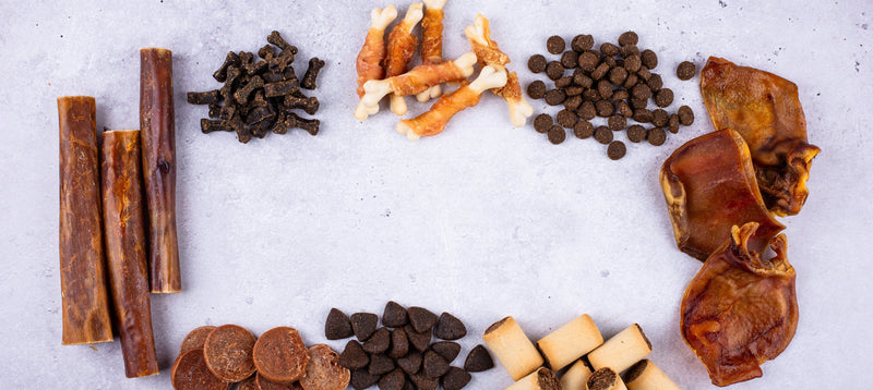 An overhead flat lay of assorted natural dog treats on a grey surface, including dried chews, skins and biscuits